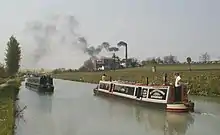 Une section droite du canal, avec deux bateaux se croisant au premier plan, avec un grand bâtiment en briques et une grande cheminée, au milieu du plan, sur la rive droite du canal. De la fumée s'échappe de la cheminée, vers le canal situé sur la gauche.