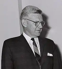 photographie en noir et blanc d'un homme blanc aux cheveux clairs portant des lunettes, une veste sombre, une chemise blanche et une cravate.