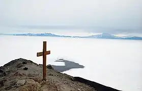 Grande croix de bois planté dans le roc au sommet d'une colline enneigée dominant le paysage ; le ciel est bleu, à l'horizon se dessine un chaîne de montagnes