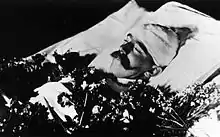 La tête entourée de bandages, laissant voir son visage portant une moustache, Rodolphe gît dans un lit la poitrine recouverte de fleurs.