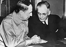 Photo en noir en blanc du général MacArthur en uniforme militaire et du premier ministre australien John Curtin( en costume sombre assis à une table. Curtin se penche vers MacArthur afin de l'écouter.
