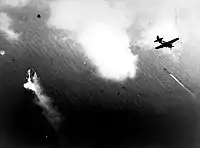Photographie en noir et blanc prise d'un avion à moyenne altitude. Un autre avion précède celui d'où a été pris la photo et un navire est visible.