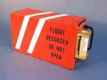 Photo couleur. Boîte rectangulaire rouge avec deux bandes réfléchissantes banches et une inscription en anglais « enregistreur de vol, ne pas ouvrir ».