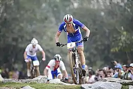  Homme à VTT portant une tenue bleu sombre, avec en arrière-plan deux poursuivants et un public.