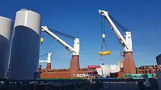 À l'arrière plan, la grue du navire de transport vient de déposer la turbine sur son ber. La grue est encore équipée du palonnier. Au premier plan de gauche à droite on voit des sections des piles des futurs éoliennes, et des bers vide pour en accueillir de nouveaux. Au premier plan au milieu à gauche on distingue une plateforme automotrice (verte) qui sert à déplacer les éléments, et à droite à l'arrière plan une des grues mobiles (verte) qui permet de manipuler les éléments... Cette grue mobile n'est pas suffisante pour déplacer les piles ou les turbines, une grande structure porteuse (non photographiée ici) sert à redresser les piles, qui arrivent horizontales par bateau, et sont redressées avant d'être stockées verticales.