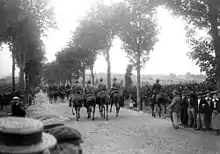 Colonne de cavaliers français sur quatre de front, de dos, la route pavée bordée par des civils.
