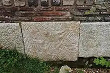 Présentation de la partie basse d'un mur avec les fondations en pierres de taille dont une est sculptée, et les niveaux supérieurs en briques et moellons.