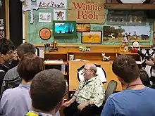 Homme âgé assis parlant avec animation à des jeunes debout autour de lui dans une pièce de travail où sont exposées des images du film