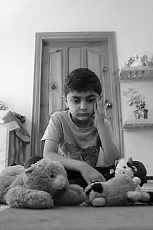 Photo en noir et blanc d'un enfant malheureux, assis dans une chambre parmi ses peluches.