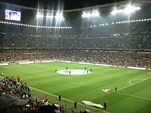 Photographie d'un grand stade de football illuminé avec des tribunes pleines.