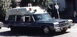 La photo est centrée sur une ambulance de la marque Cadillac de 1973. Le bas de la voiture, les portes, le capot sont de couleur bleu tandis que le haut de la voiture est blanc.