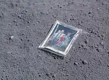 Photographie en couleur d'une photo de famille de Charles Duke posée sur le sol lunaire. La photo est sous emballage plastique.