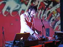 Photo en vue de trois-quart de Daedelus. Daedelus est un homme aux cheveux noirs, avec de larges rouflaquettes. Il porte un costume blanc et une cravate bleue. Il sourit devant du matériel audio et un ordinateur portable floqué du logo d'Apple. Le fond est rouge-mauve et on devine le mot « Daedelus » écrit en larges lettres blanches.