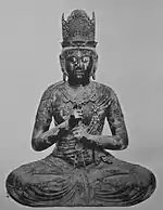 Vue de face d'une statue représentée portant une couronne et montrant la Vajra Mudrā, enfermant l'index de la main gauche avec le poing de la main droite.