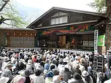 Photo couleur montrant des spectateurs assis devant une scène ouverte sur laquelle se joue une pièce de kabuki.