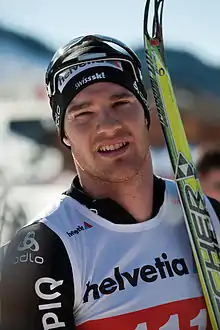 Portait d'un homme, des lunettes de ski sur sa tête, tenant ses skis sur sa gauche.