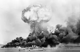Plan large en noir et blanc d'un nuage de fumée résultant d'une explosion. On peut voir un navire de guerre au premier plan.