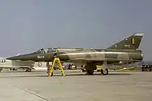 Un Mirage 5 immobile au sol, sur un tarmac, avec d'autres avions stationnés à l'arrière-plan.