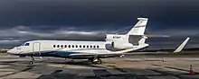 Un Falcon 7X garé au parking, vu de profil. L'avion blanc présente une peinture bleue sur le bas de la carlingue, séparée du blanc par des liserés gris. L'avion est immatriculé N73KT. L'avion est éclairé par la lumière du Soleil et mis en évidence par le contraste avec les nuages sombres à l'arrière-plan.