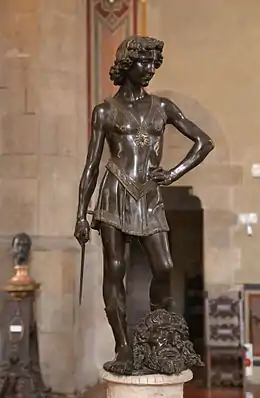 Photographie de la statue de bronze d'un jeune homme vêtu de vêtements légers tenant dans sa main droite une épée courte la main gauche sur la hanche. Debout, dans une posture triomphante et détendue entre ses pieds est posée sur une tête coupée.