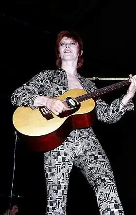 Un homme aux cheveux rouges en combinaison blanche et noire tenant une guitare.