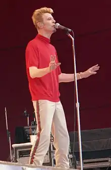 Photo d'un homme debout sur une scène en train de chanter dans un micro qui porte un pantalon blanc et un t-shirt rouge