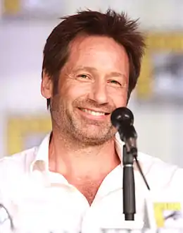 Homme brun souriant habillé d'une chemise claire, derrière un microphone noir.