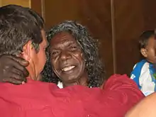 Gulpilil est au centre de l'image, souriant ; un homme habillé en rouge tourne le dos à l'objectif ; les deux hommes ont un bras sur l'épaule de l'autre.