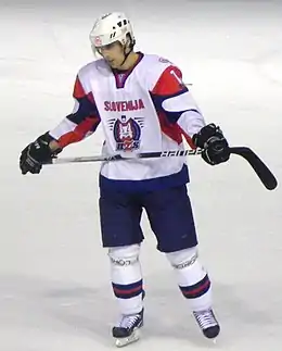 Photographie couleur d’un hockeyeur, de buste, légèrement de profil, le regard dans le vague, portant un maillot blanc avec les épaules vertes claires