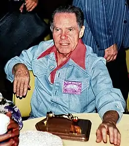 Photographie d'un homme âgé assis devant une table. Il porte une veste bleue.