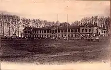 Photographie en noir et blanc imprimé en sépia sur carte postale. De jeunes gens jouent sur un terrain de rugby avec un bâtiment qui domine le terrain, derrière les poteaux de rugby.