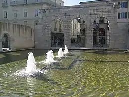 Entourés de bâtiments de style gallo-romain, un bassin rectangulaire laisse jaillir quatre jets d'eau chaude au centre d'une retenue d'eau.