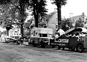 noir et blanc, chars rangés sur le côté gauche d’une rue ;  parfois publicitaires (marque de cigarettes) ; arbres bordant l’avenue, magasins sur le côté ; slogans publicitaires peu visibles