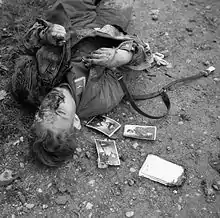 Photo noir et blanc prise le 23 décembre 1943 à Ortona, en Italie. Dans la partie gauche de la photo, le cadavre d’un soldat allemand gît (tête vers le bas de la photo) sur un sol de terre pierreuse. Trois photos de soldats sont visibles près de son épaule droite.