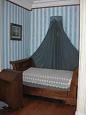 Photo d'un lit bateau de teinte acajou, recouvert d'un couvre-lit en dentelles blanches et surmonté d'un dais de velours gris, dans une petite chambre tapissée d'un motif à larges rayures bleues et blanches.