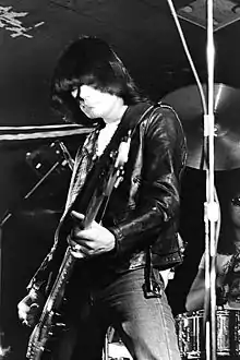 photo en noir et blanc d'un homme vêtu d'un jean et d'un blouson noirs, une guitare à la main et devant des cymbales.