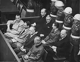 Photographie en hauteur et en noir et blanc d'un groupe d'homme en trois rangs, à gauche et au milieu assis portant des vêtements de ville, à droite debout portant l'uniforme et des casques blancs.
