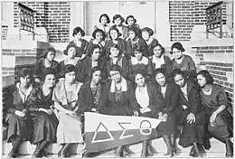 Un groupe de jeunes femmes afro-américaines, en uniforme, assises sur les escaliers d'un bâtiment en brique. Le rang de devant tient une banderole à l'effigie de la sororité.