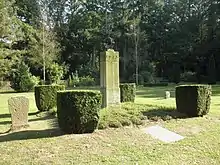 Monument en pierre, entouré de quatre buissons, au milieu d'une surface d'herbe, dans un cimetière.