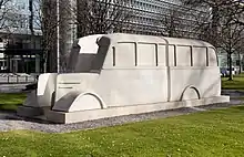 Photographie en couleur d'une sculpture en béton d'un bus gris, coupé longitudinalement.