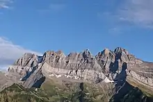 Parois rocheuses verticales en formes de dents de scie surplombant une pente verte garnie de quelques arbres.