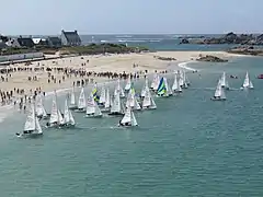 Vue montrant des bateaux à voiles quittant une plage, et une foule rassemblée pour regarder le spectacle.