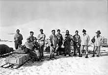 Photographie noir et blanc de 10 hommes debout dans la neige, avec du matériel et un traineau.