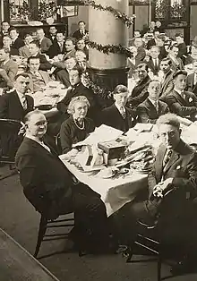 Photo noir et blanc d'un ensemble de personne autour de plusieurs tables sur lesquels se trouvent des cadeaux de Noël déballés. La salle est ornée de guirlandes et autres décoration de Noël.