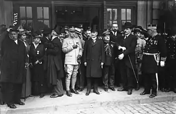 Photo en noir et blanc de plusieurs personnes (civils et militaires) se tenant debout ; l’homme au centre, moustachu, porte une longue veste et un haut-de-forme