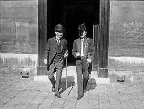 Photo en noir et blanc de deux hommes en train de marcher, épée de l’Académie française à la main : celui de droite a une moustache blanche, un costume trois pièces avec chemise à faux col et chapeau melon ; l’autre, moustache foncée, porte l’habit vert de l’Académie