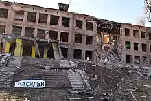 Photographie d'un immeuble en briques de trois étages éventré, fenêtres manquantes, avec des débris jonchant le sol et un fragment de panneau portant l'inscription « Василькі[в] ».