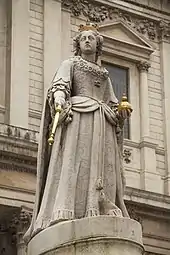 Photo d'une statue d'une femme en pierre grise, à l'exception des insignes de la royauté (couronne, sceptre, orbe) qui sont dorés