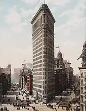 Photographie d'un immeuble très effilé de forme triangulaire d'une vingtaine d'étages surplombant les bâtiments voisins et les rues en contrebas où circulent des calèches et des tramways.