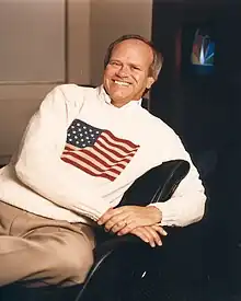 Photographie d'un homme accoudé à sa chaise de bureau et portant un pull blanc avec le drapeau américain en son centre.